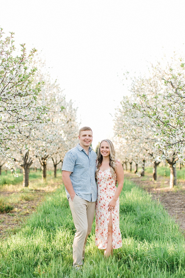 A couple stands smiling in a cherry orchard in spring in Traverse City, Michigan