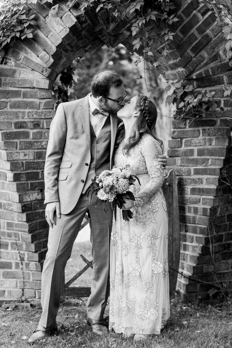 This is a black and white photo of a bride and groom kissing under an arch. 