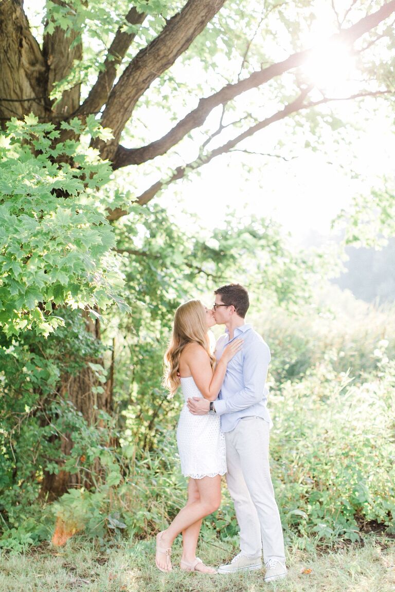 This is an engaged couple kissing under a tree in the summer at The Village at Grand Traverse Commons