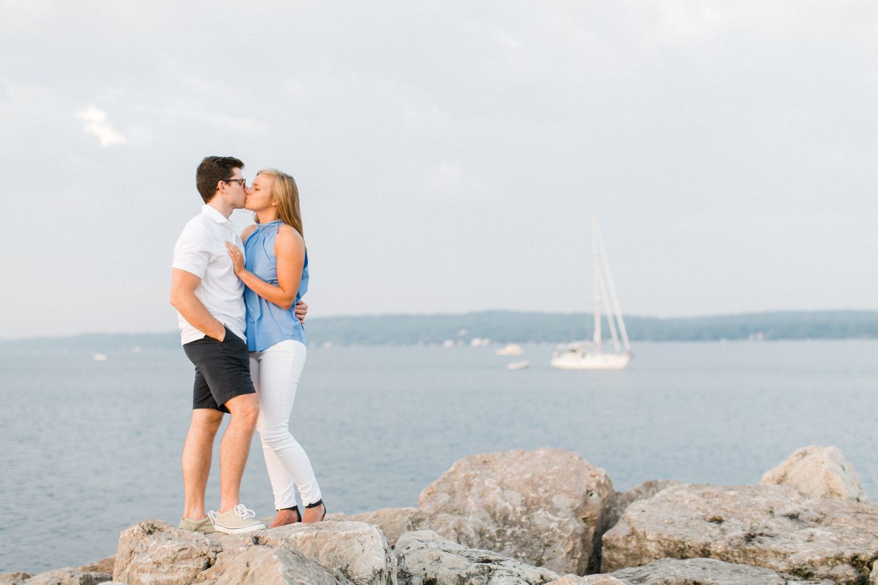 This is an engaged couple kissing on a rock by the water in Traverse City, Michigan
