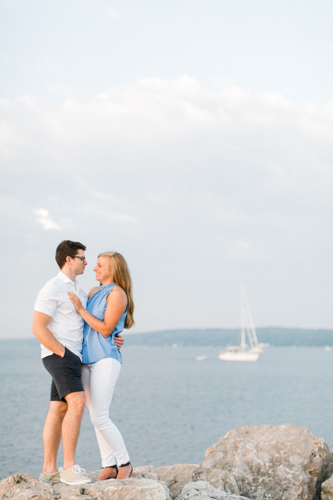 This is an engaged couple on a rock, looking at each other, with a sailboat in the background on Grand Traverse Bay in Traverse City, Michigan
