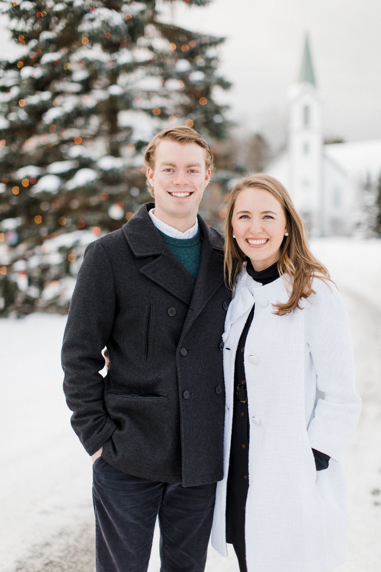 This is an engaged couple smiling in Downtown Harbor Springs with a Christmas tree and church in the background
