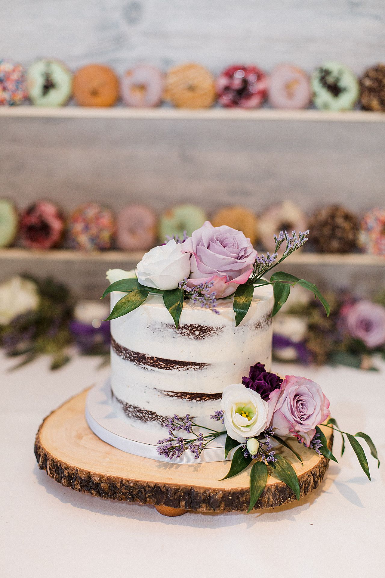 A single tier wedding cake with white frosting, purple flowers sitting on a wooden slab with donuts in the background