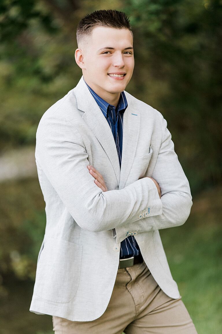 This is a photo of a male senior portrait with a tan blazer on in a wooded area in Traverse City, Michigan