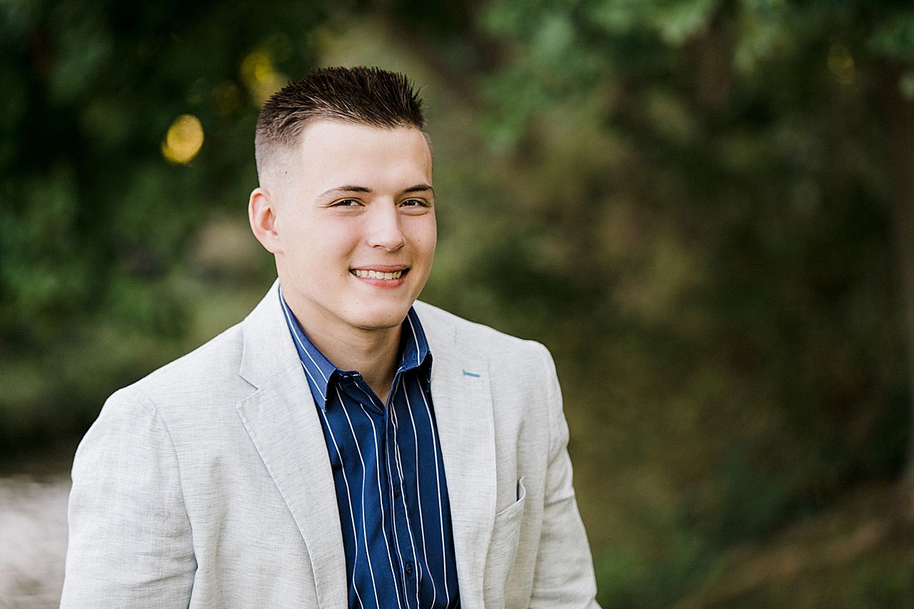 This is a photo of a male senior portrait with a tan blazer on in a wooded area in Michigan