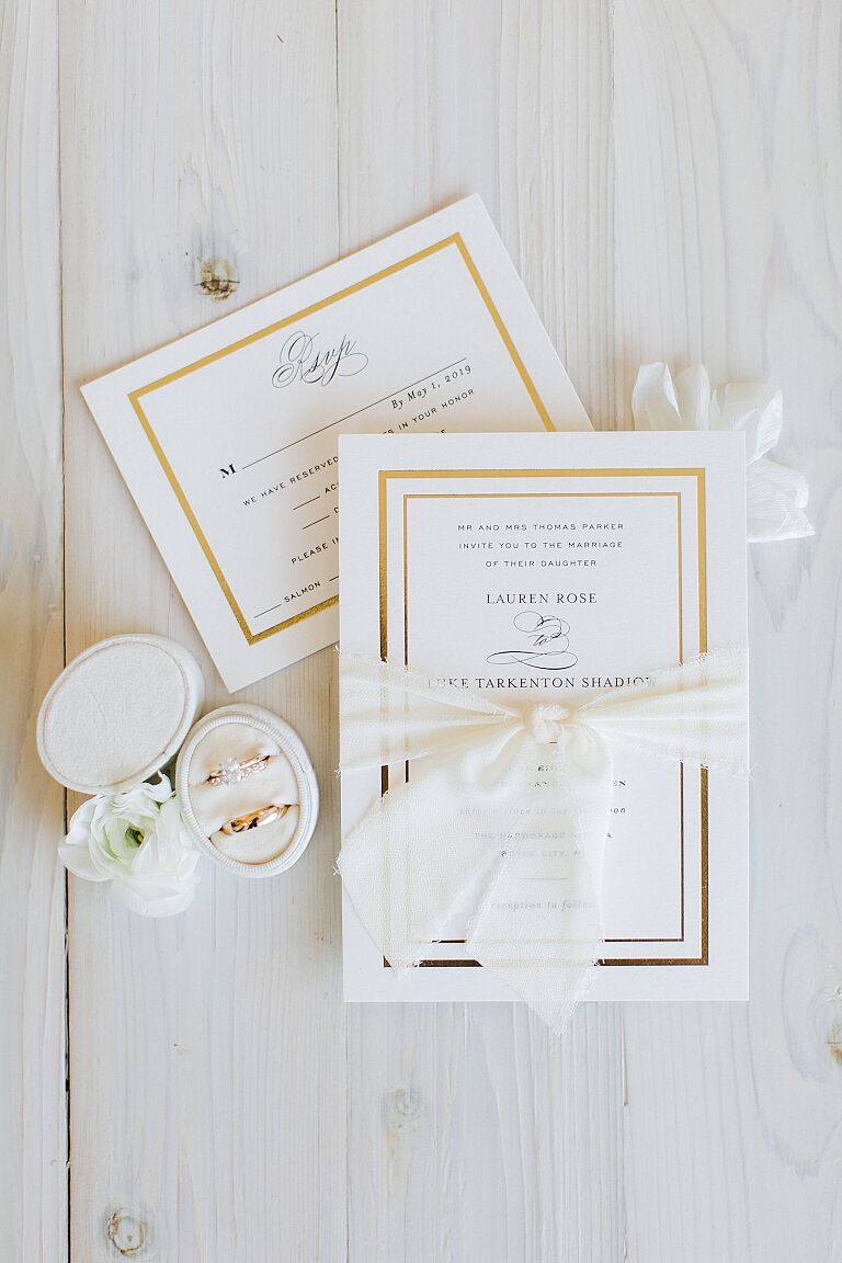 White and gold wedding invitation with a white ribbon tied around it with wedding rings to the side