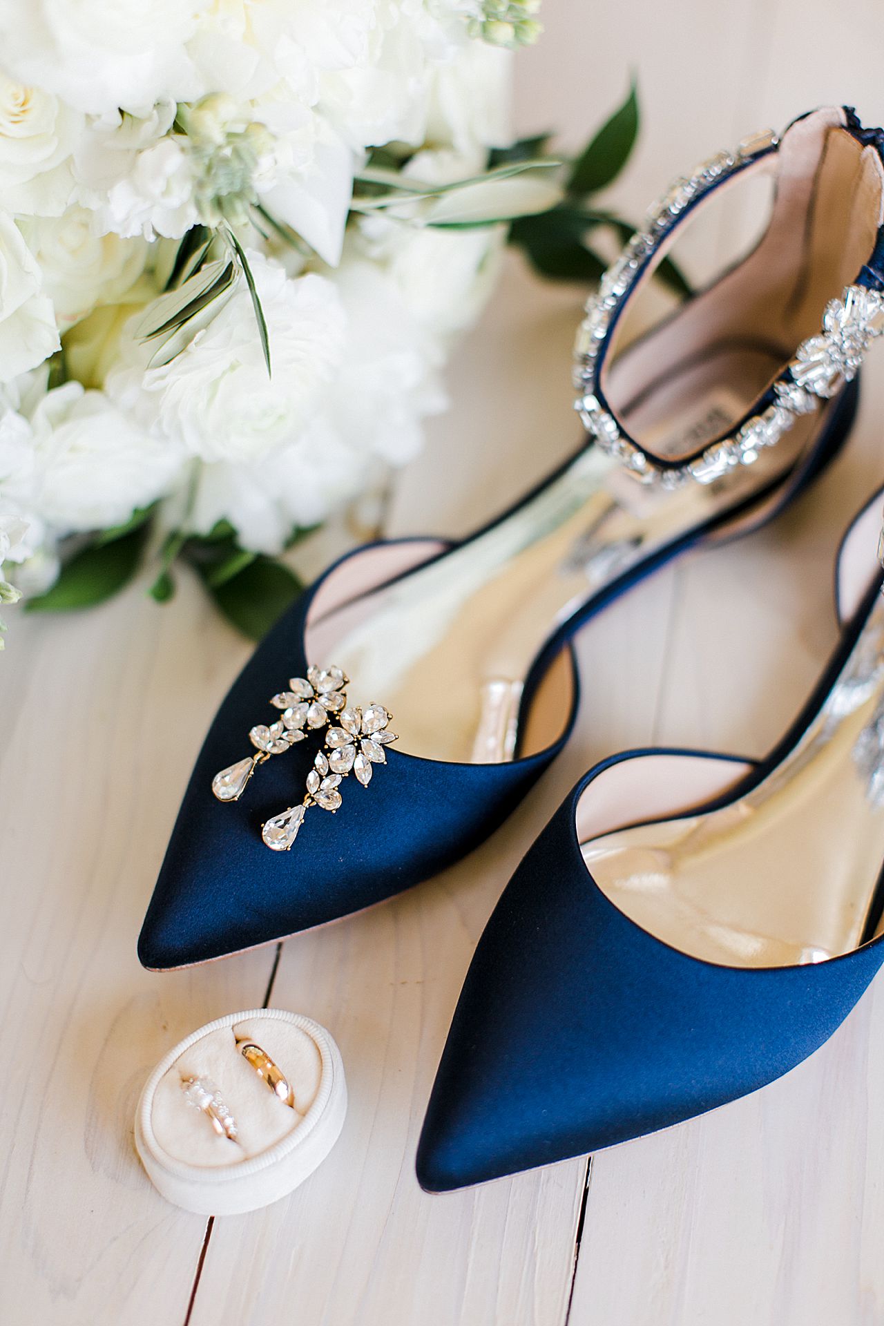 A pair of long crystal earrings rested on a pair of navy blue Badgley Mischka women's Mary Jane shoes with a white bridal bouquet in the background and wedding rings in the foreground