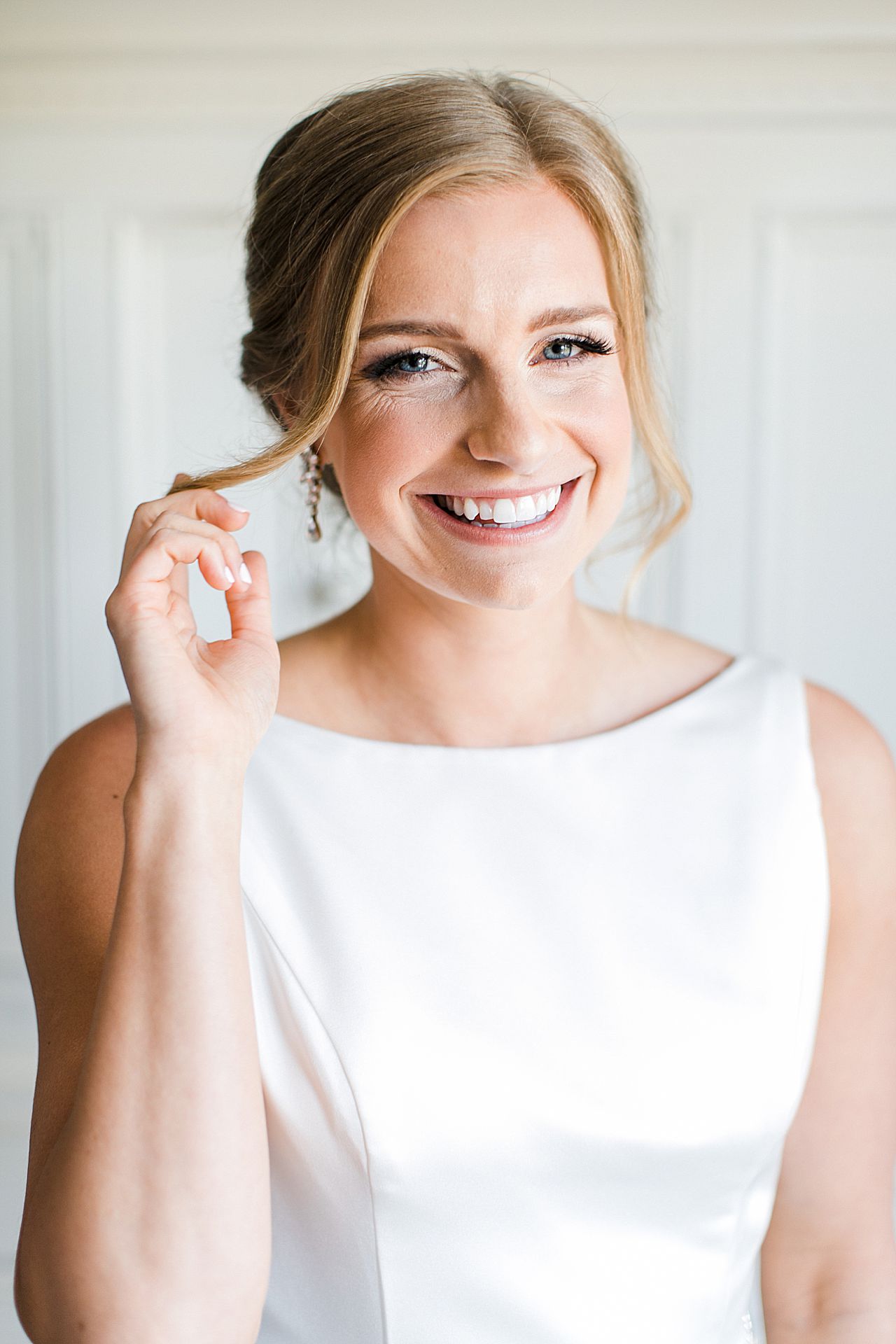 A bride in her wedding dress looking into the camera, smiling and pushing back a small piece of hair to the side of her face