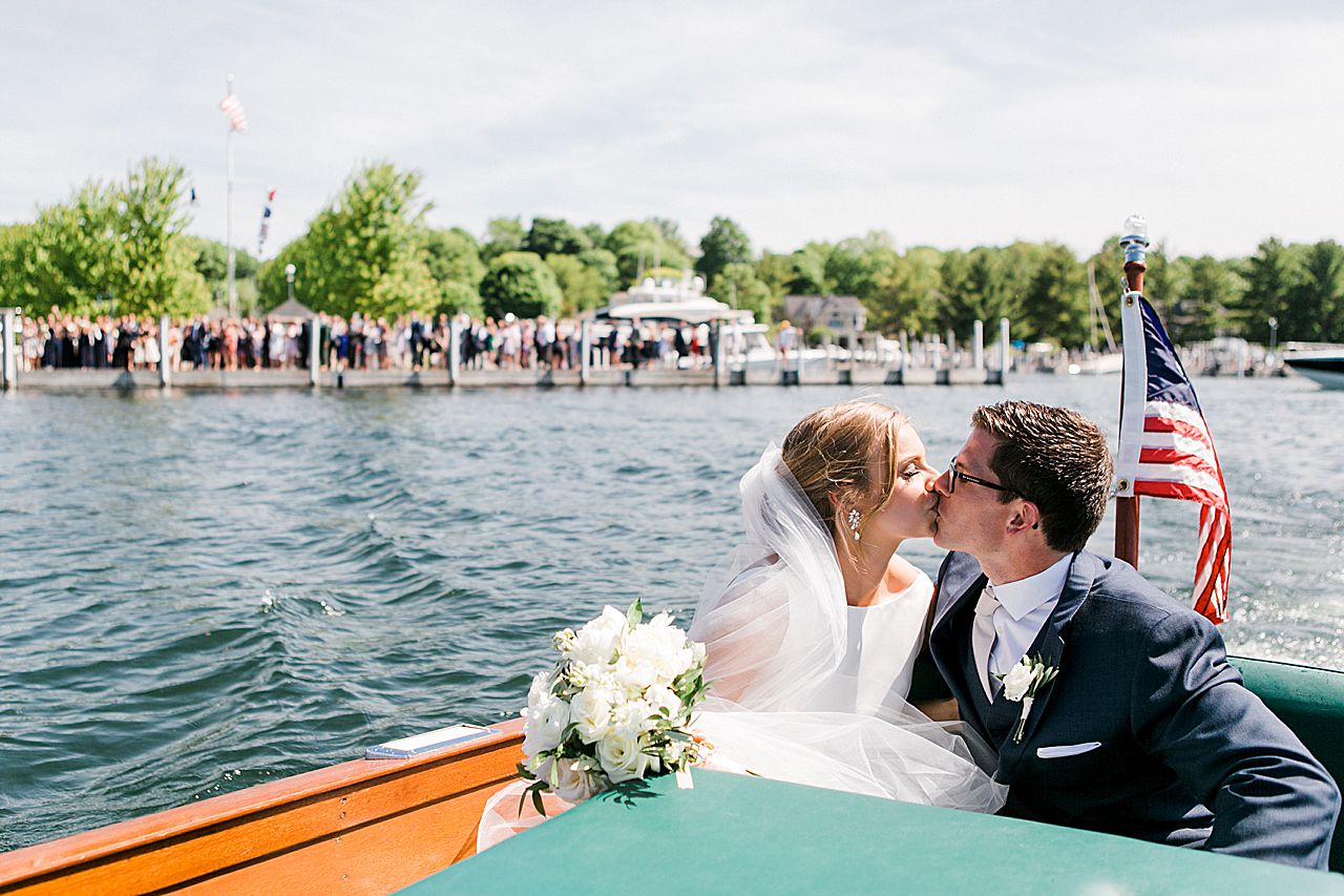 Bride and Groom kissing on a Chris Craft boat on Lake Charlevoix with wedding guests in the background on the dock