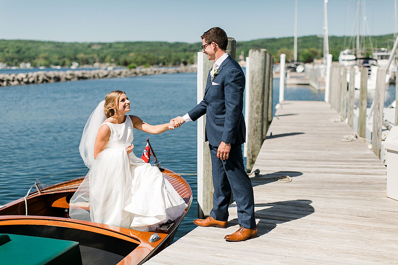 Groom holding the bride's hand as she steps out of a Chris Craft boat onto the dock at The Harborage Marina in Boyne City, Michigan