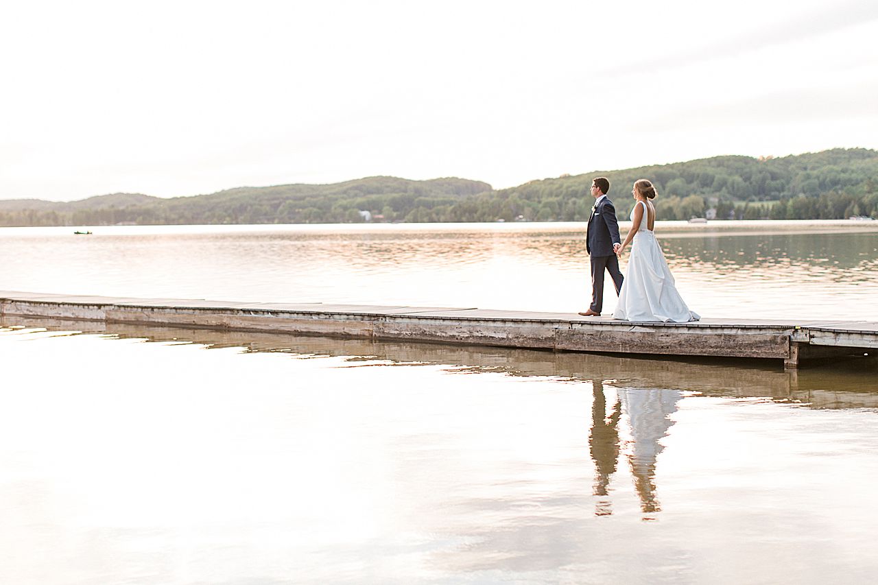 Bride and groom walking on a dock on Deer Lake with their reflection in the water in Boyne Falls, Michigan