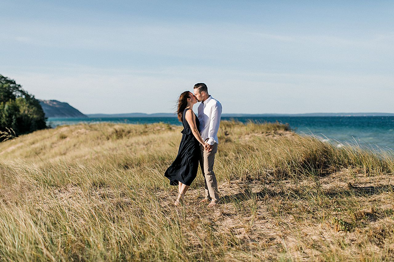 This is an engaged couple holding hands and kissing on top of a sand dune along Sleeping Bear Dunes National Lakeshore