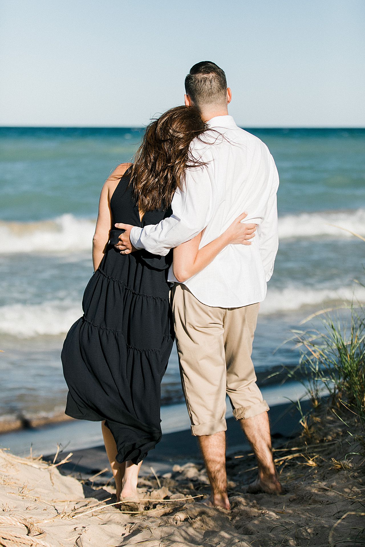 This is an engaged couple with their arms around each other looking at the vibrant blue water along Sleeping Bear Dunes National Lakeshore