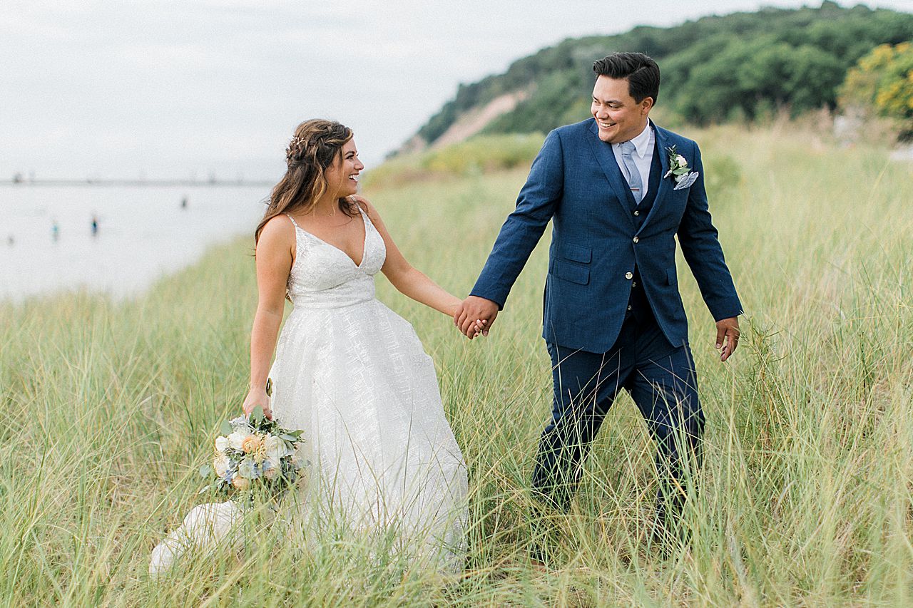 A bride and groom smiling and walking in dune grass along Lake Michigan in Frankfort, Michigan