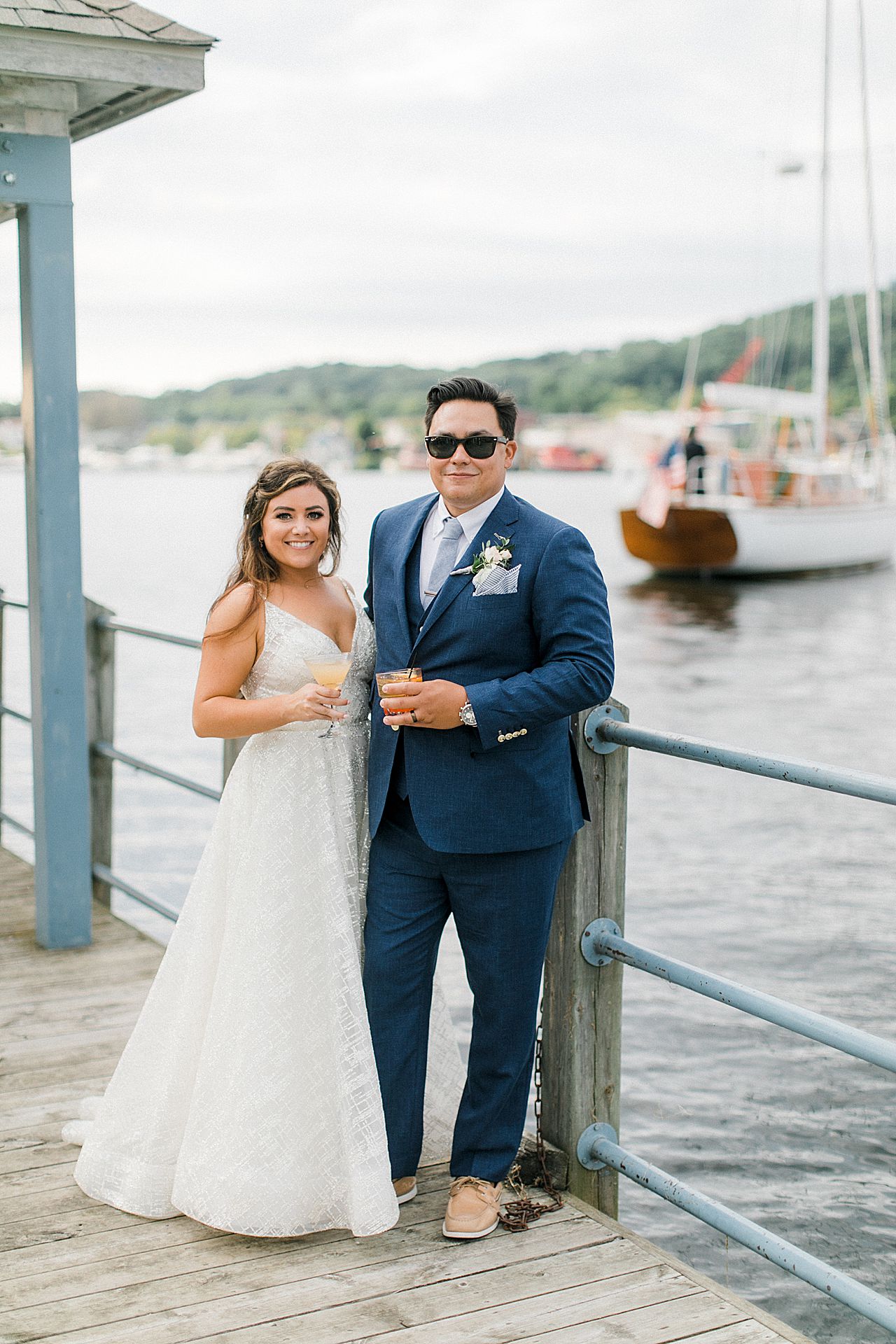 A bride and groom holding their signature wedding cocktails with a sailboat in the background in Elberta, Michigan