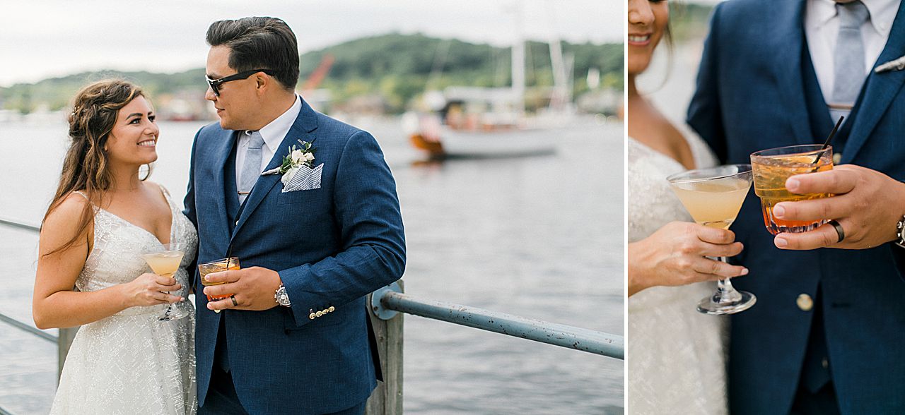 A bride and groom holding their signature wedding cocktails with a sailboat in the background in Elberta, Michigan