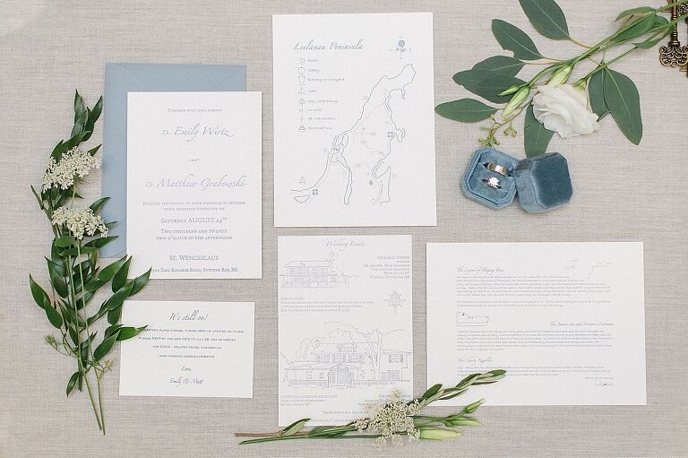 A light blue and white wedding stationery set on a light grey background with green and white florals