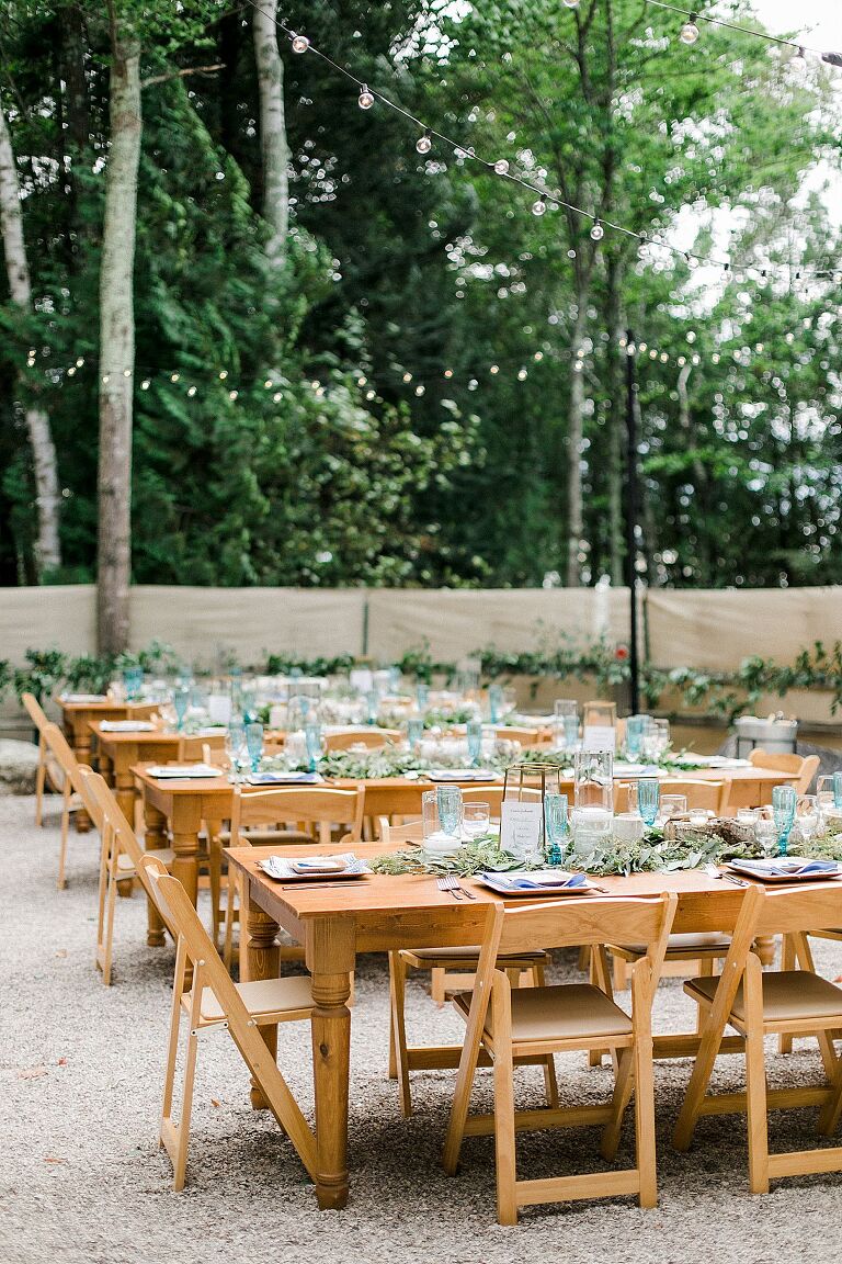Long wooden reception tables and chairs with greenery, lanterns, candles, and blue accents
