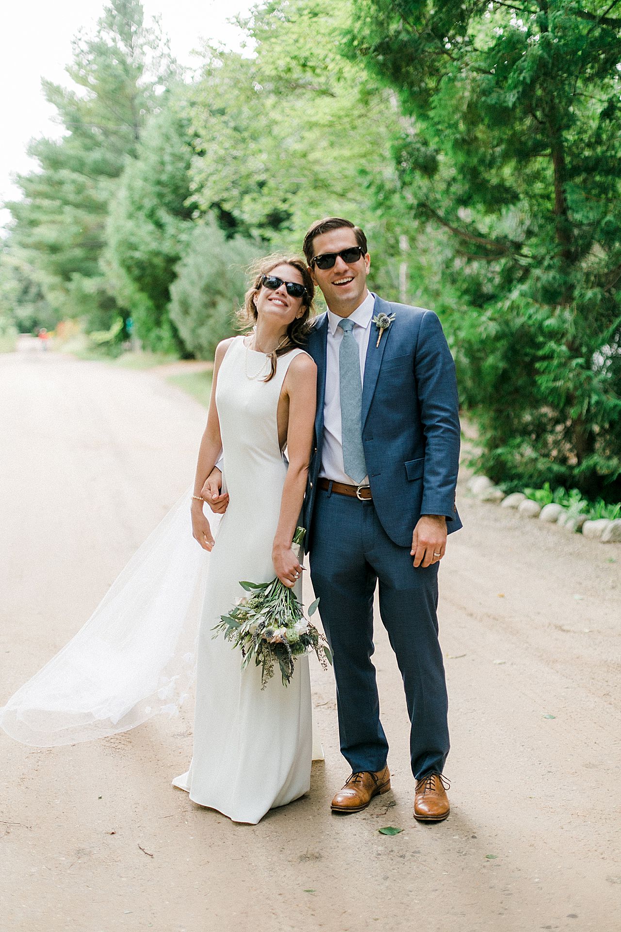 A bride and groom portrait of them smiling and wearing their sunglasses on a dirt road