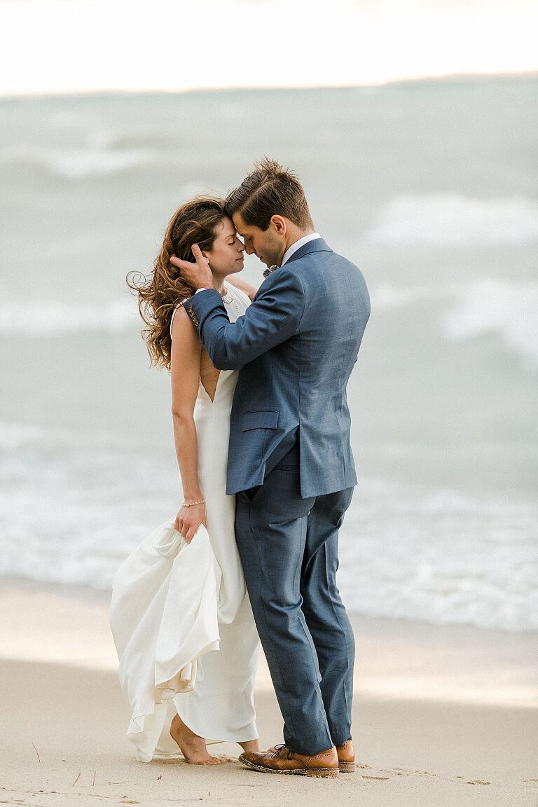A bride and groom with their eyes closed, foreheads touching, and wind blowing through their hair on Lake Michigan