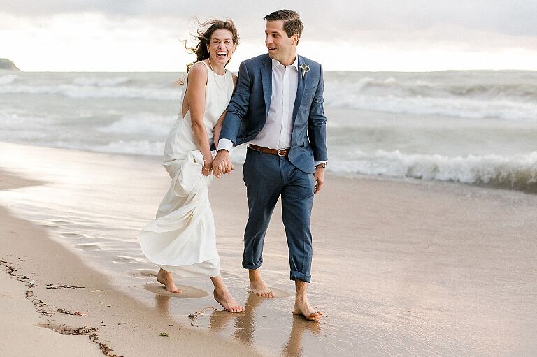 A bride laughing while walking down the lakeshore with her groom while holding hands