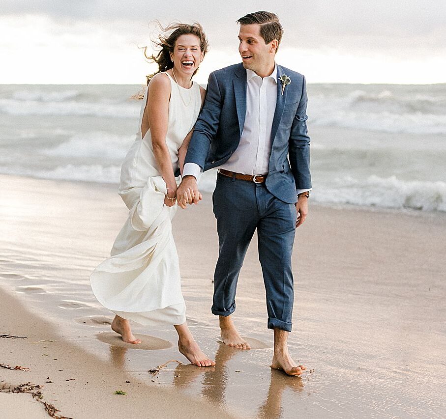 A bride laughing while walking down the lakeshore with her groom while holding hands
