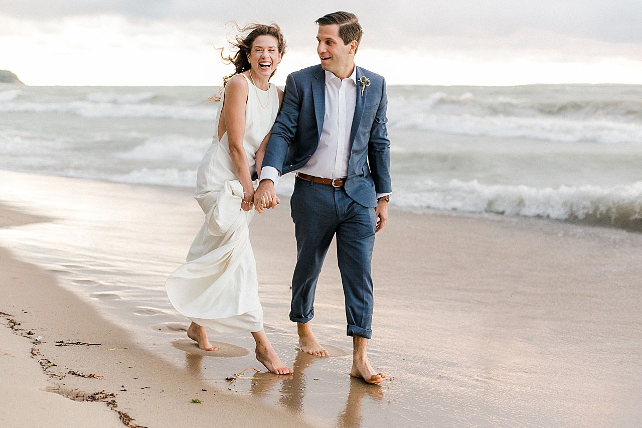 A bride laughing while walking down the lakeshore with her groom while holding hands