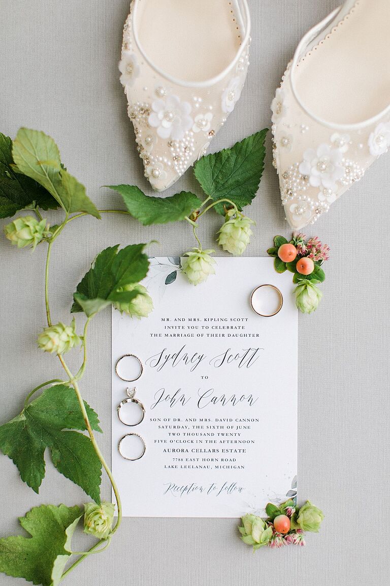 A wedding invitation, white shoes, and rings with hops and flowers around