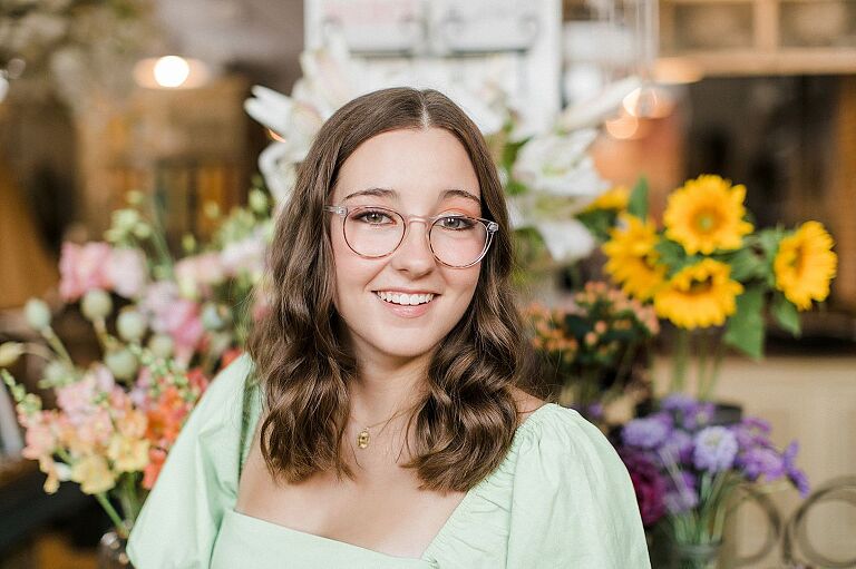 A senior portrait session in Charlevoix, Michigan with a girl wearing a green dress and glasses in a floral shop