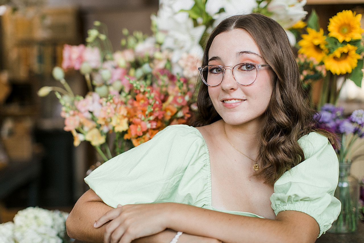 A senior portrait session in Northern Michigan with a girl wearing a green dress and glasses in a floral shop