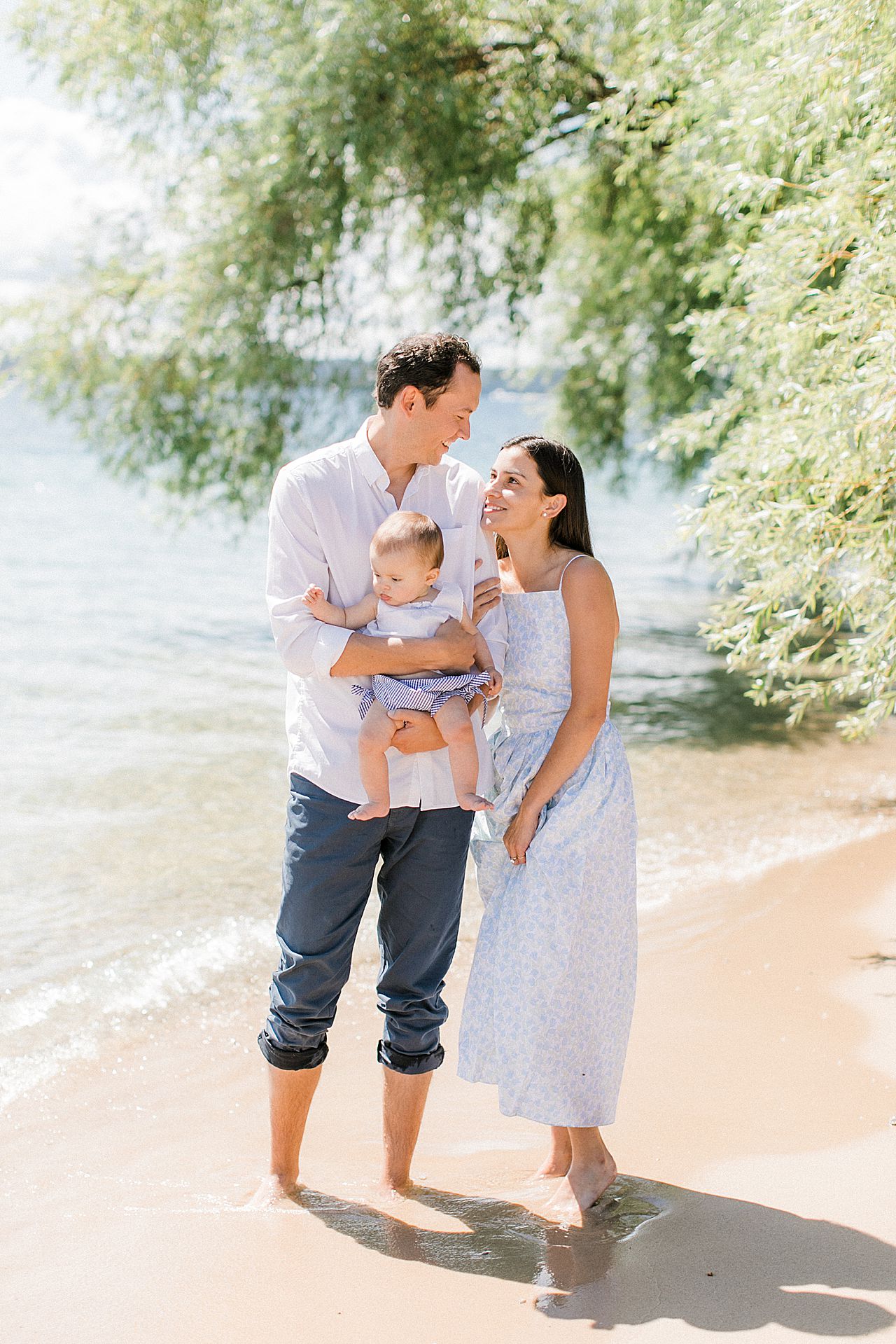 A mother and father looking at each other while holding their baby daughter on a sunny day by the lake in Northern Michigan