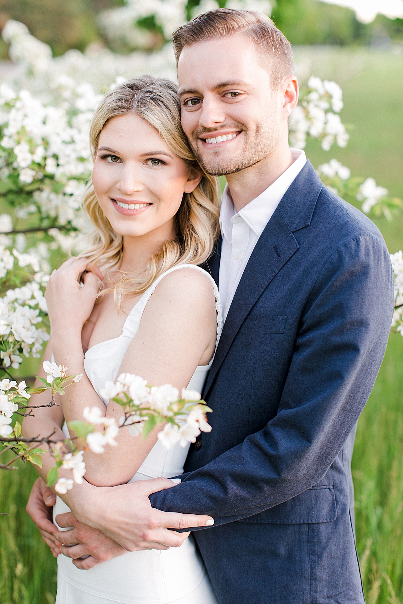 An engaged couple smiling and cuddling in a field with blooming trees on Old Mission Peninsula in Northern Michigan