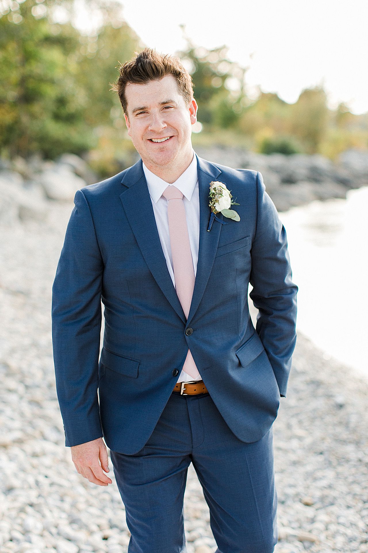 A groom in a blue suit smiling by the lake in Michigan