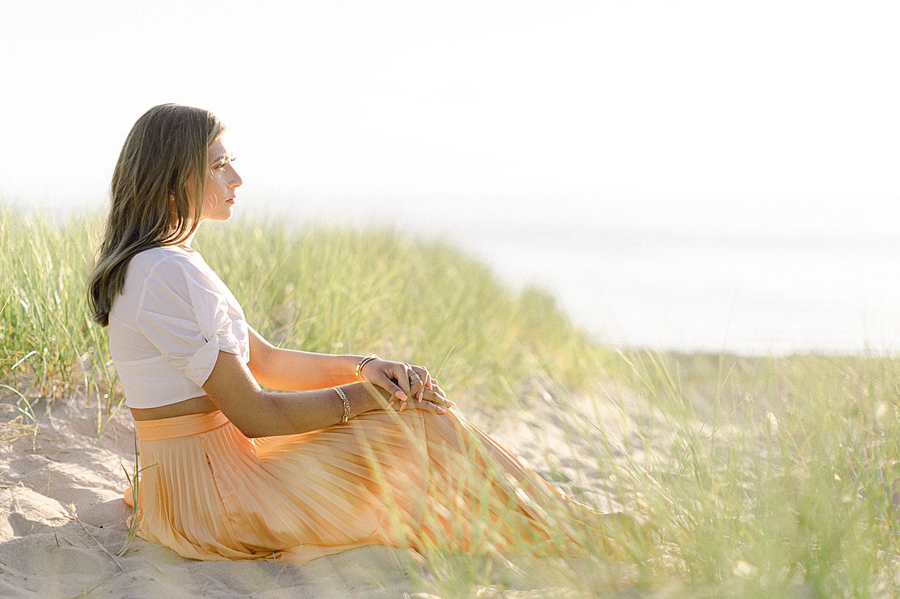 A high school senior girl sitting in the sand and looking out at Lake Michigan