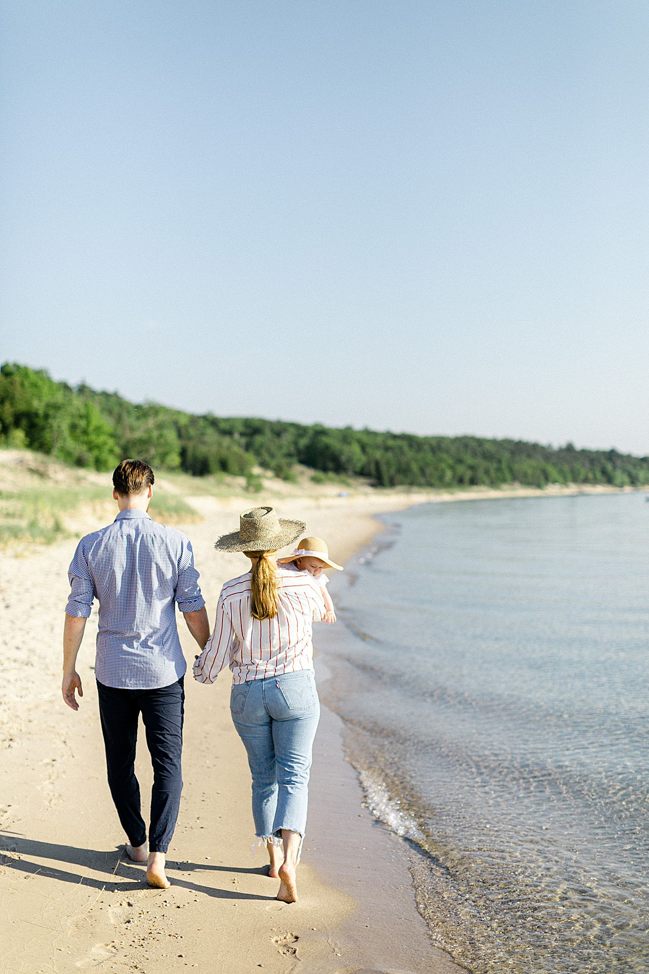 A mother and father holding hands and walking along the lake on a summer day with their daughter in Michigan