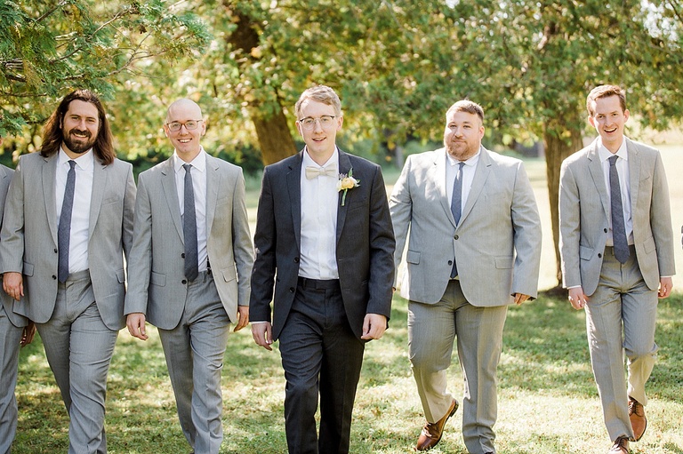 A groom and groomsmen walking and smiling in the shade of a tree on a sunny day.