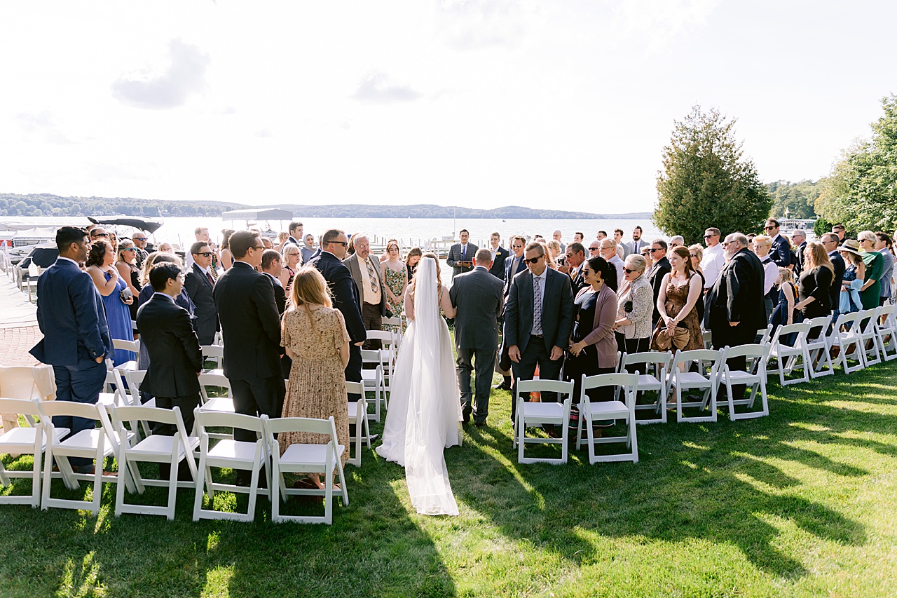 The guests of a wedding watching a bride being escorted to the groom on a lake in the sunshine.