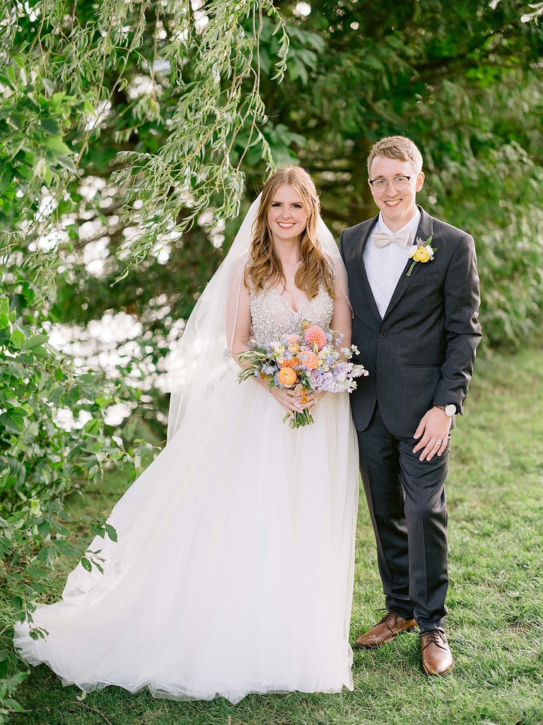 A portrait of a smiling bride and groom under a tree branch on a summer day.