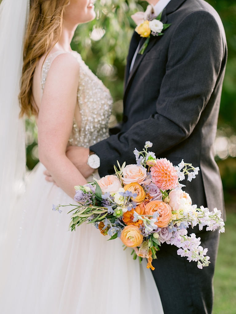 Portrait of bridal bouquet with groom holding bride in the background in the shade of trees.