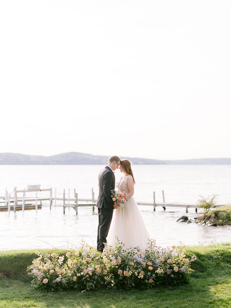 A bride and groom leaning into each other on a lake behind pastel floral ground piece.
