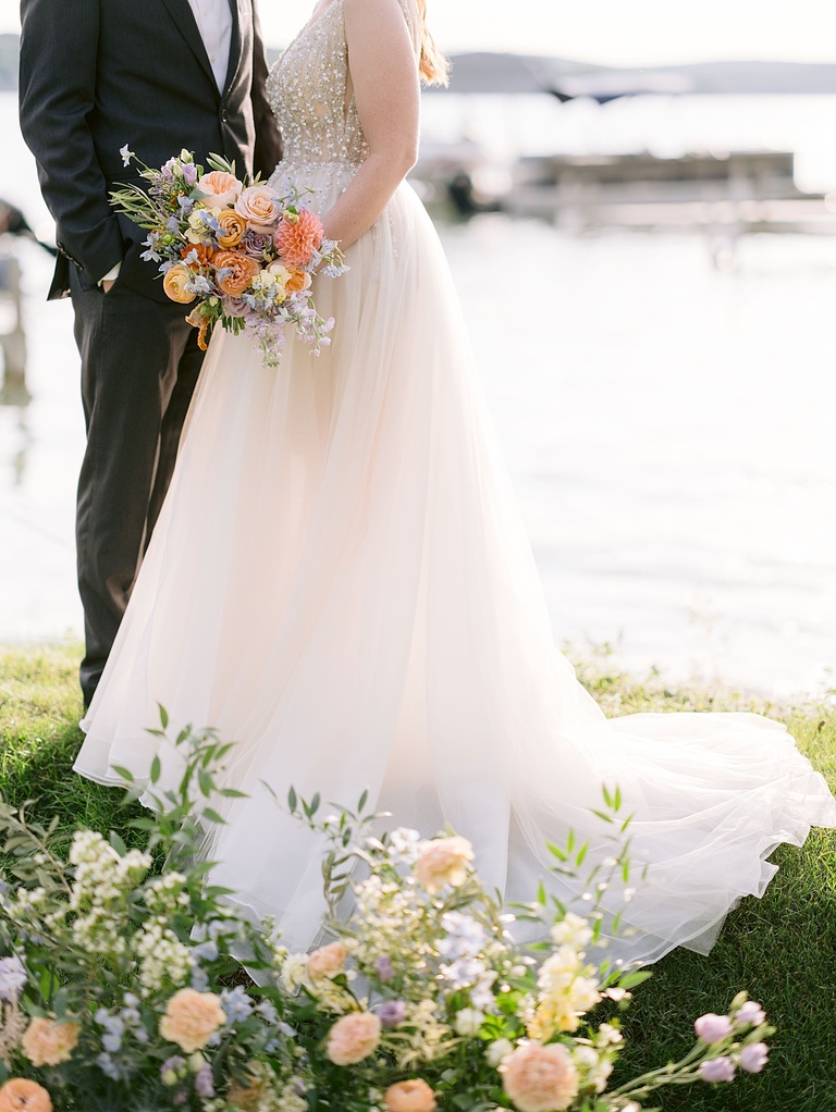Portrait of bride and groom from the grass in front of a lake to their shoulders while lit by sunlight.