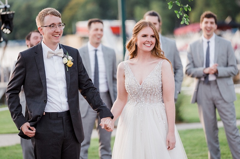 A smiling bride and groom entering their reception while groomsmen smile and life in the background.