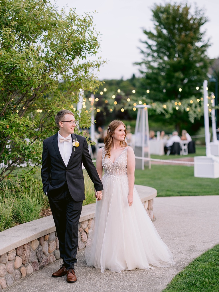 A bride and groom walking and admiring their scenery with reception party lights glow in the background.