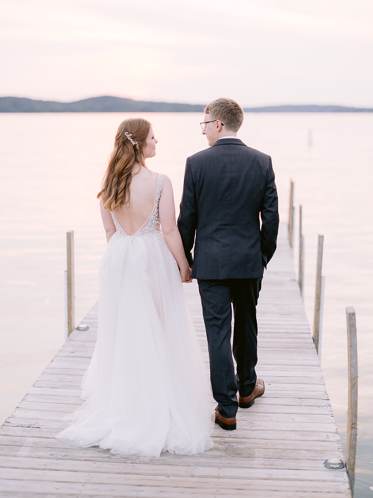 A bride and groom walking on a dock with a sunset lake and hills in the background.