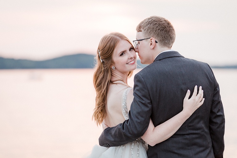A bride looks over the grooms shoulder smiling while he leans in for a kiss on a sunset lake.
