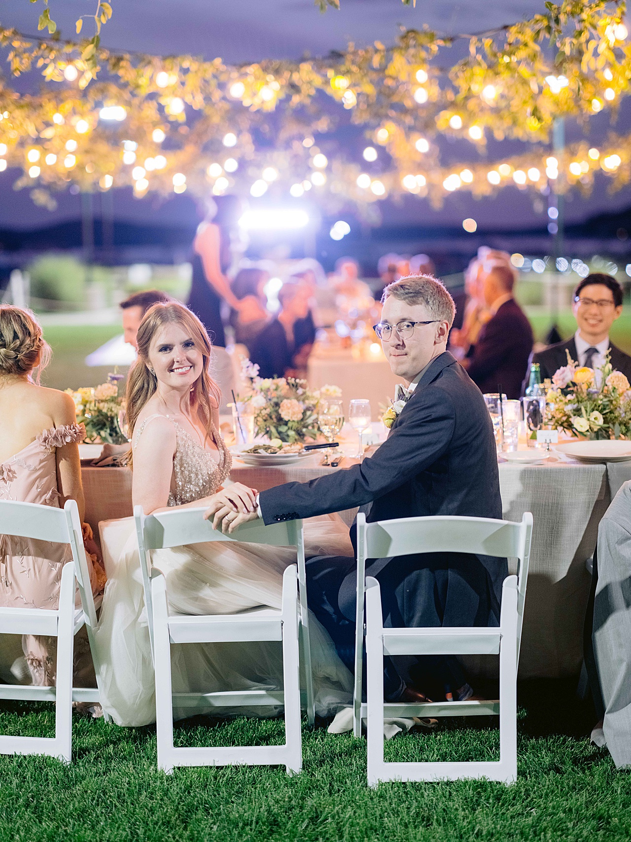 A bride and groom looking over their shoulders with outdoor reception party lights and guests in the background.