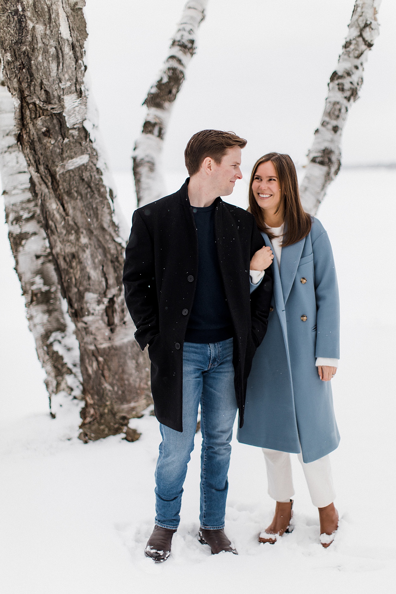 An engaged couple looking at each other in the winter with Mullett Lake and a birch tree in the background