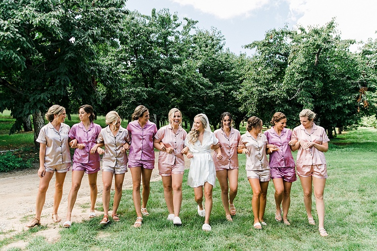A bride and her 9 bridesmaids in pink pajamas walking together in an orchard in Michigan