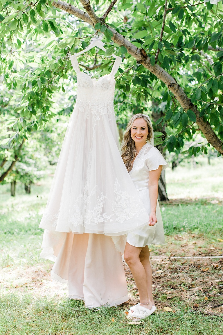 A bride standing next to her wedding dress that is hanging from a tree in an orchard in Northern Michigan