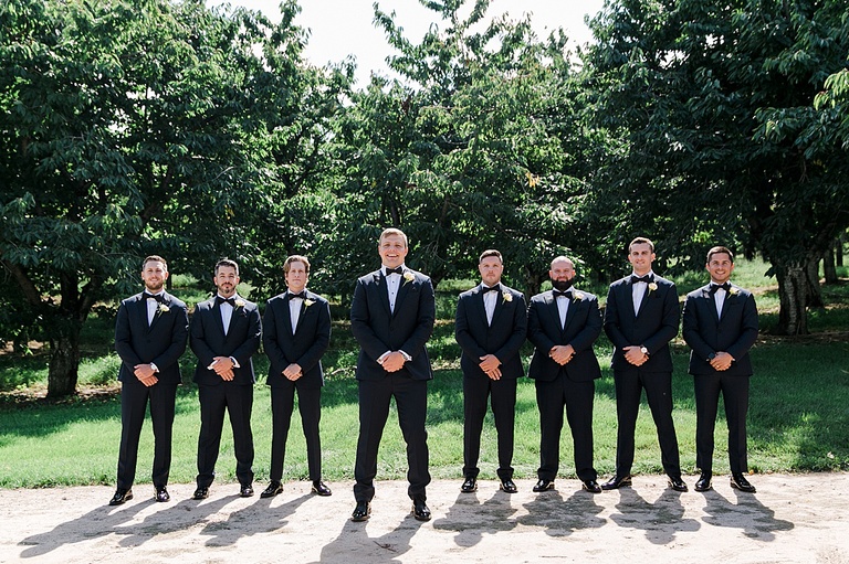 A groom and his groomsmen taking portraits on a gravel road with and orchard in the background in Michigan